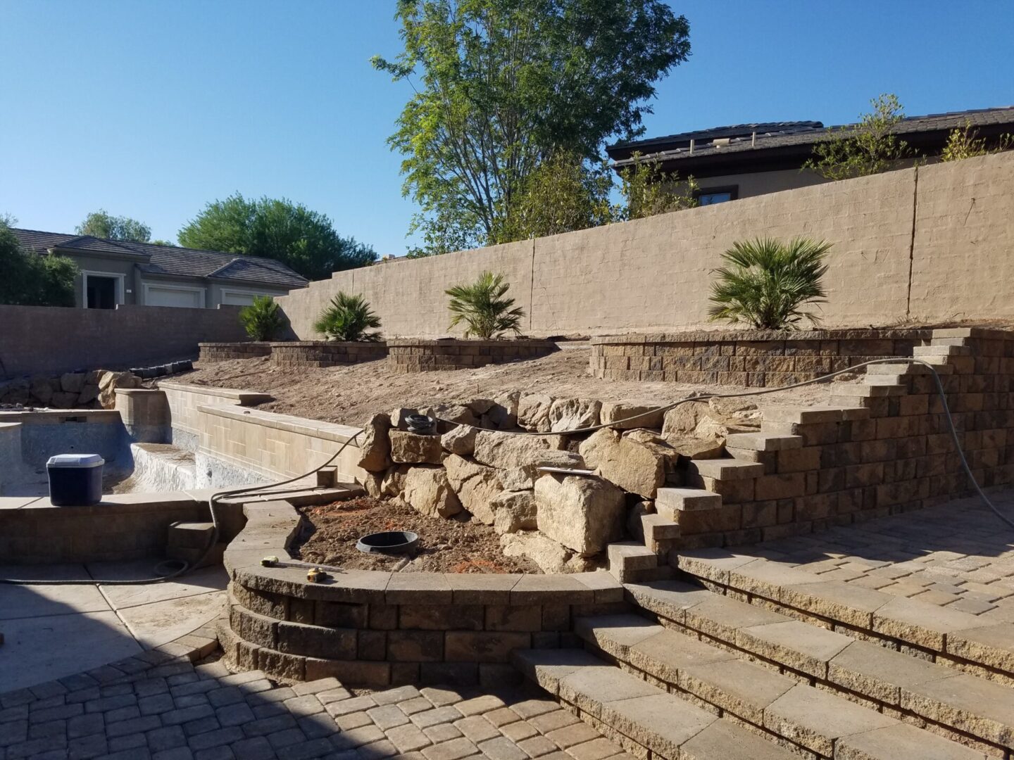 A terraced backyard with stone steps, retaining walls, and a fire pit area under clear blue sky.